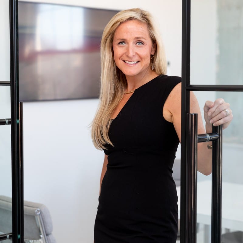A woman with long blonde hair wearing a sleeveless black dress stands in a modern office, smiling and holding open a glass door. A blurred TV screen and office furniture are visible in the background.