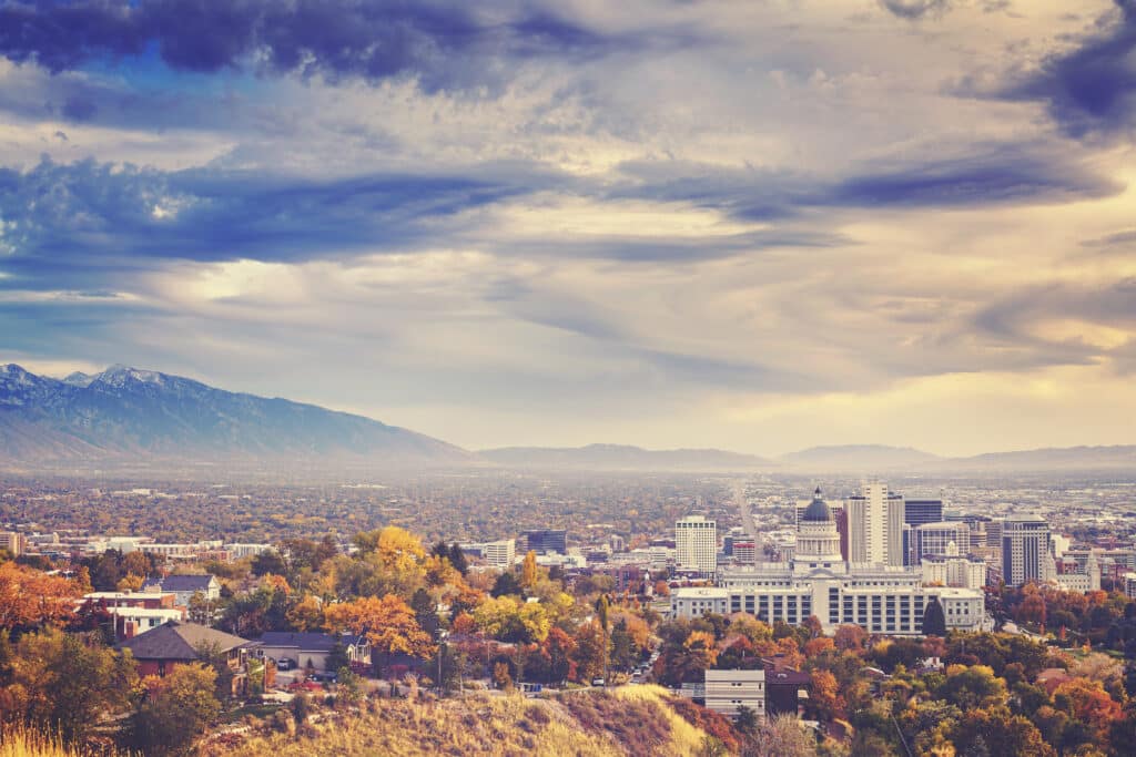 A scenic view of a city with tall buildings surrounded by autumn-colored trees, mountains in the background, and a dramatic, cloudy sky overhead.
