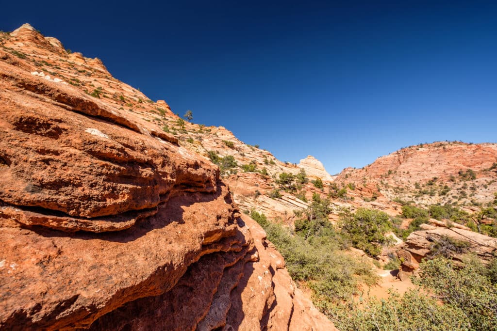 Red rock formations and rugged cliffs under a clear blue sky, with sparse greenery scattered among the rocky landscape, possibly in a desert or canyon area.