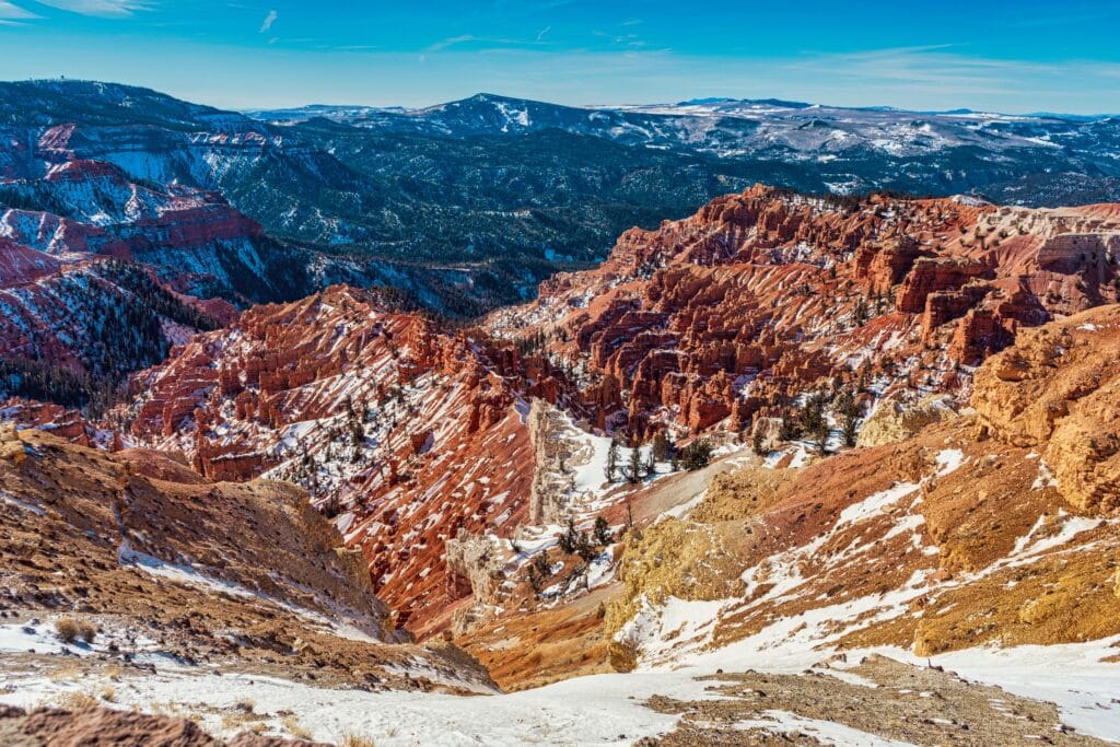 A scenic view of a rugged canyon with vibrant red rock formations dusted with snow, set against distant forested mountains and a bright blue sky.