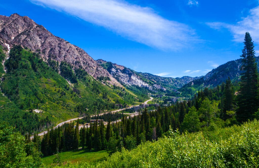 A scenic mountain valley with rugged peaks, lush green trees, and a winding road running through the landscape under a bright blue sky with wispy clouds.