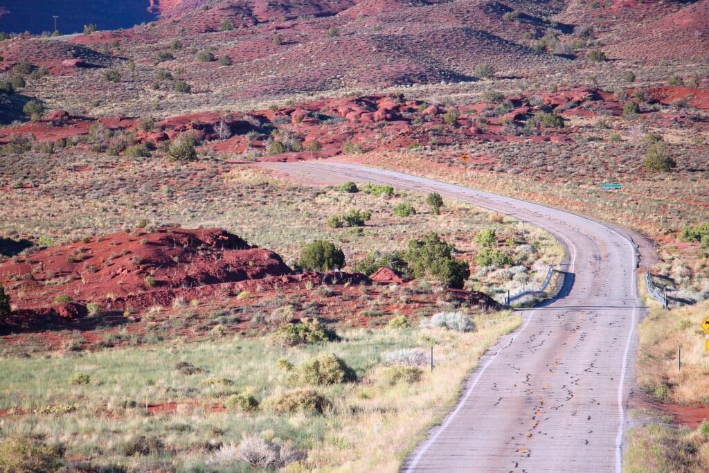 A winding paved road cuts through a dry, red and green desert landscape with low hills and sparse vegetation under clear daylight.