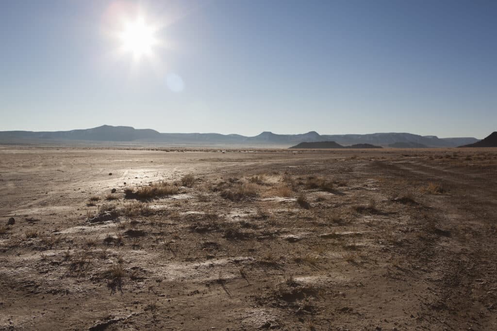 A dry, sunlit desert landscape with sparse vegetation, rough ground, and distant low mountains under a clear blue sky. The bright sun is high in the sky, casting long shadows across the scene.
