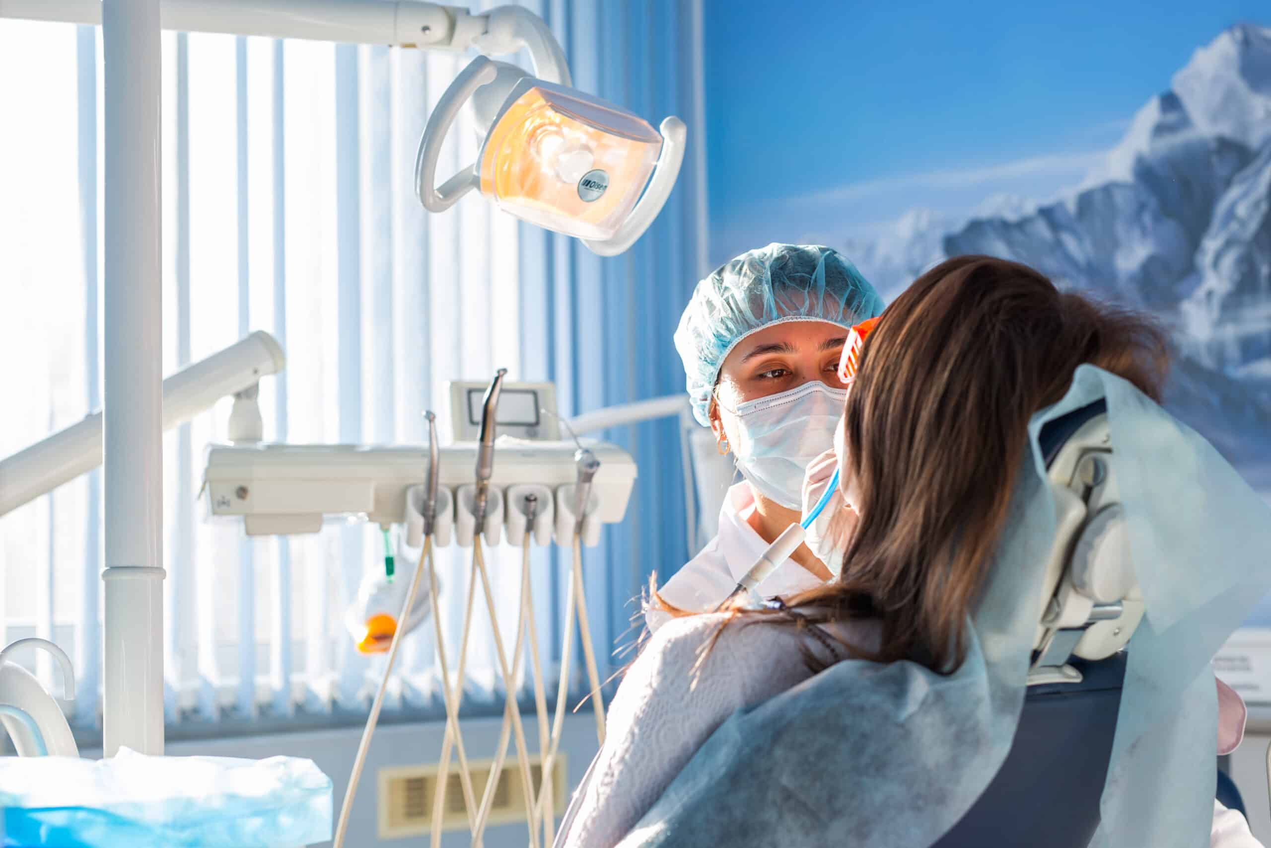 A dentist wearing a mask and cap examines a patient sitting in a dental chair under a bright overhead light in a modern dental clinic.