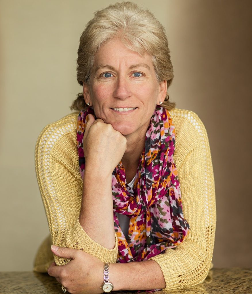 A smiling middle-aged woman with short blonde hair, wearing a yellow sweater and a colorful scarf, rests her chin on her hand while leaning on a table.
