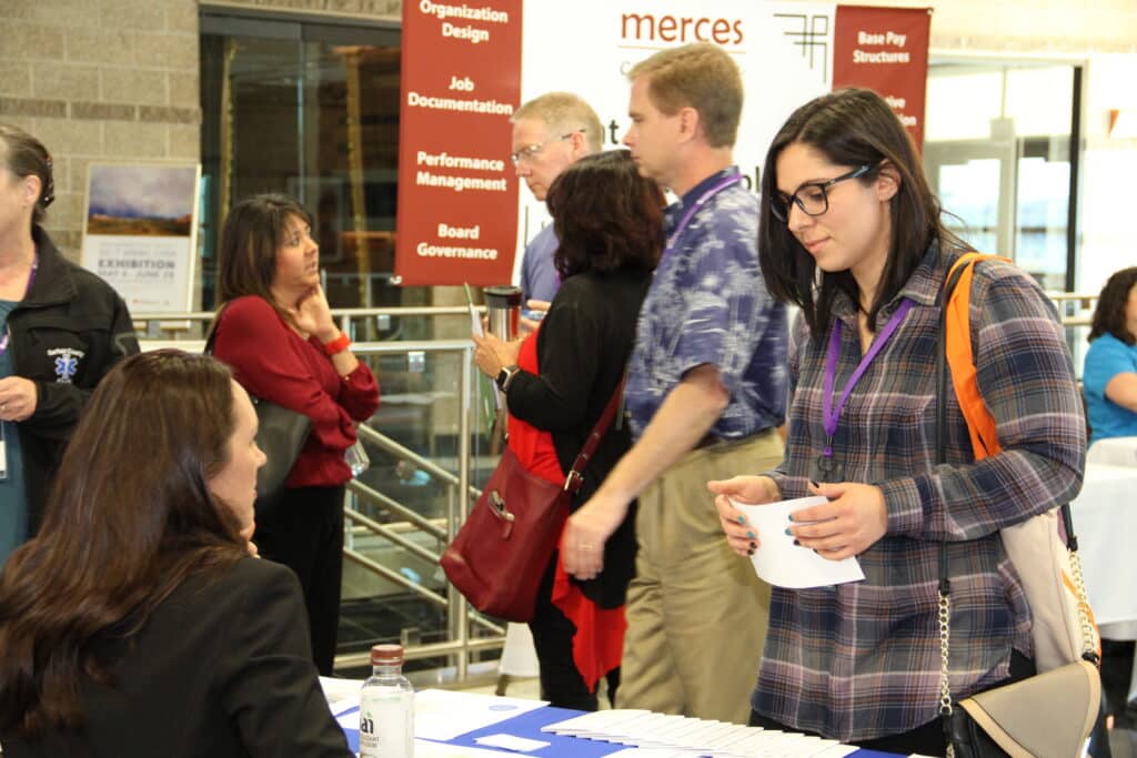 A woman speaks to a recruiter at a job fair booth, while other attendees wait in line. Informational signs about job documentation and governance are visible in the background.