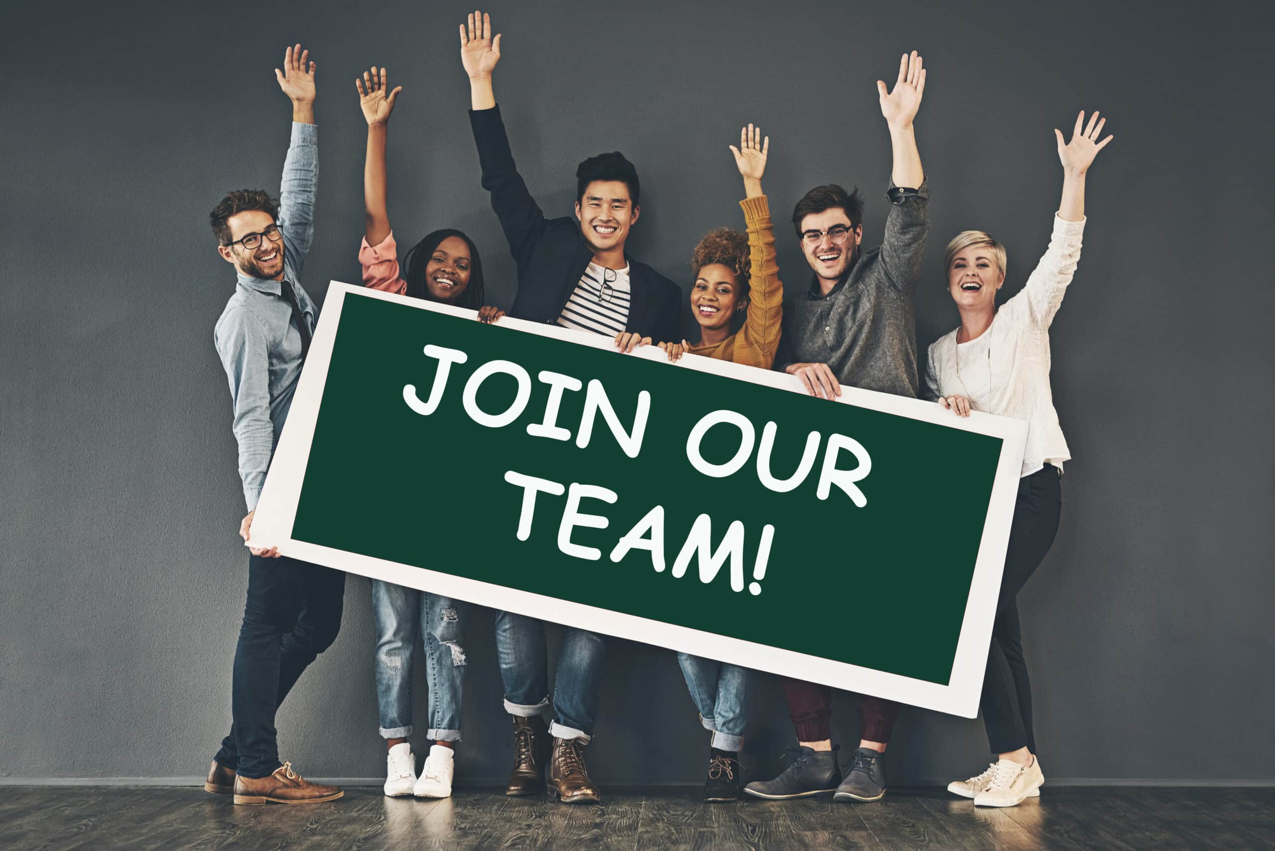 Six smiling people stand together raising their hands, holding a large green sign that reads JOIN OUR TEAM! in bold white letters, against a plain wall background.