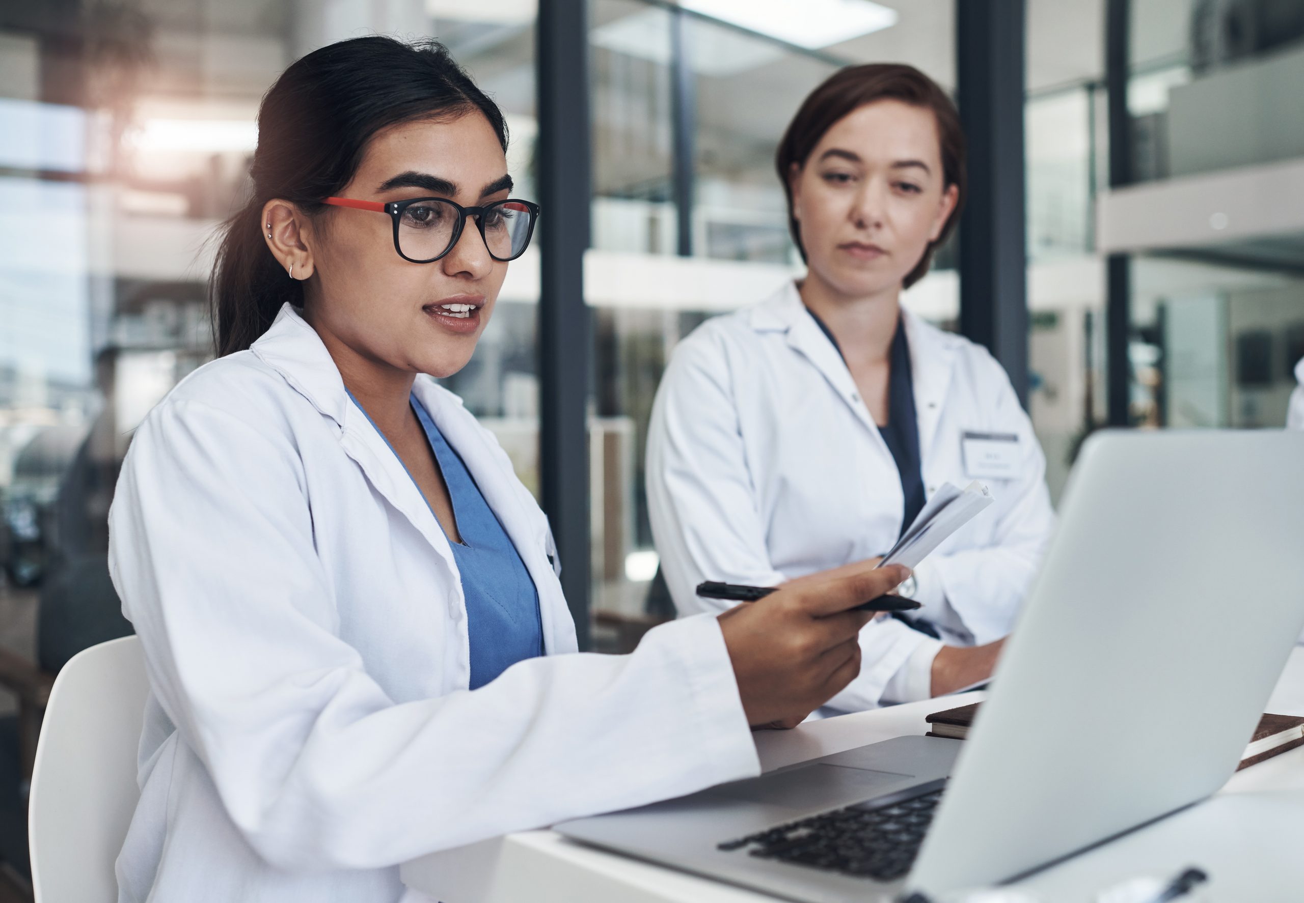 Two women in white lab coats sit at a desk with a laptop. One speaks while holding papers and a pen, and the other listens. They appear to be discussing medical information in a modern office setting.